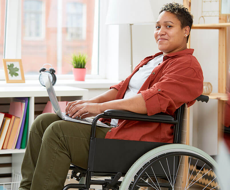 A woman in a wheelchair types on a laptop and smiles toward the camera.
