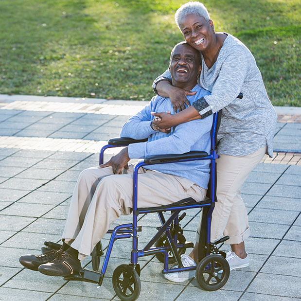 A woman hugs a man in a wheelchair in a park.