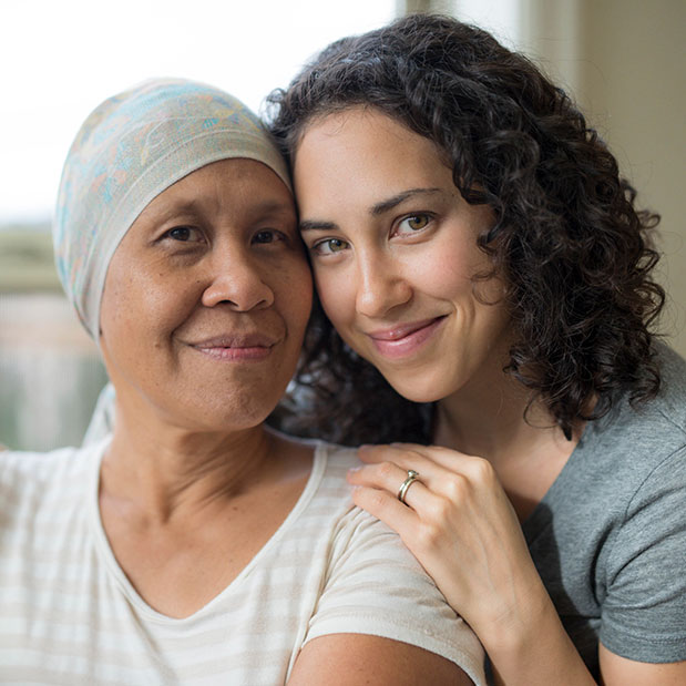 A younger woman rests her hands on the shoulders of an older woman and they smile toward the camera.