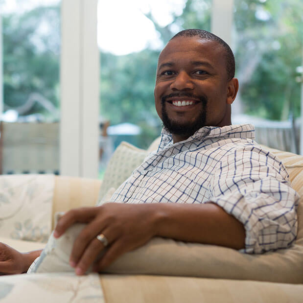 A man sits on a couch with a laptop, smiling toward the camera.
